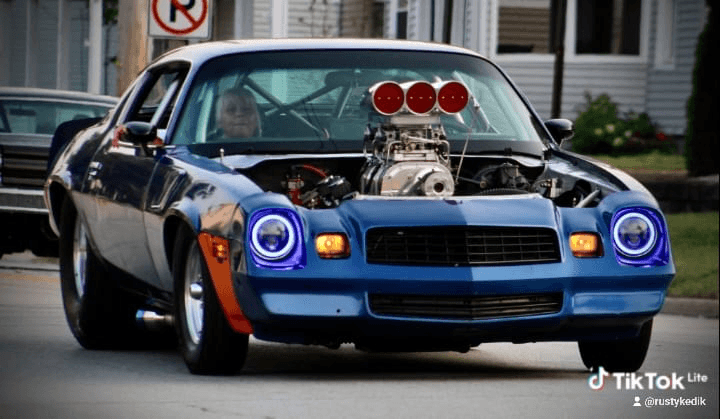 Blue second-gen Camaro with a supercharger driving on the street, halo headlights glowing and roll cage visible inside.