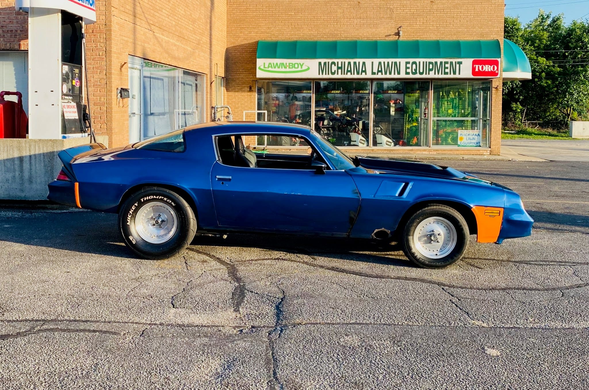 Blue second-gen Camaro parked in front of Michiana Lawn Equipment, side profile view with drag tires and hood removed.