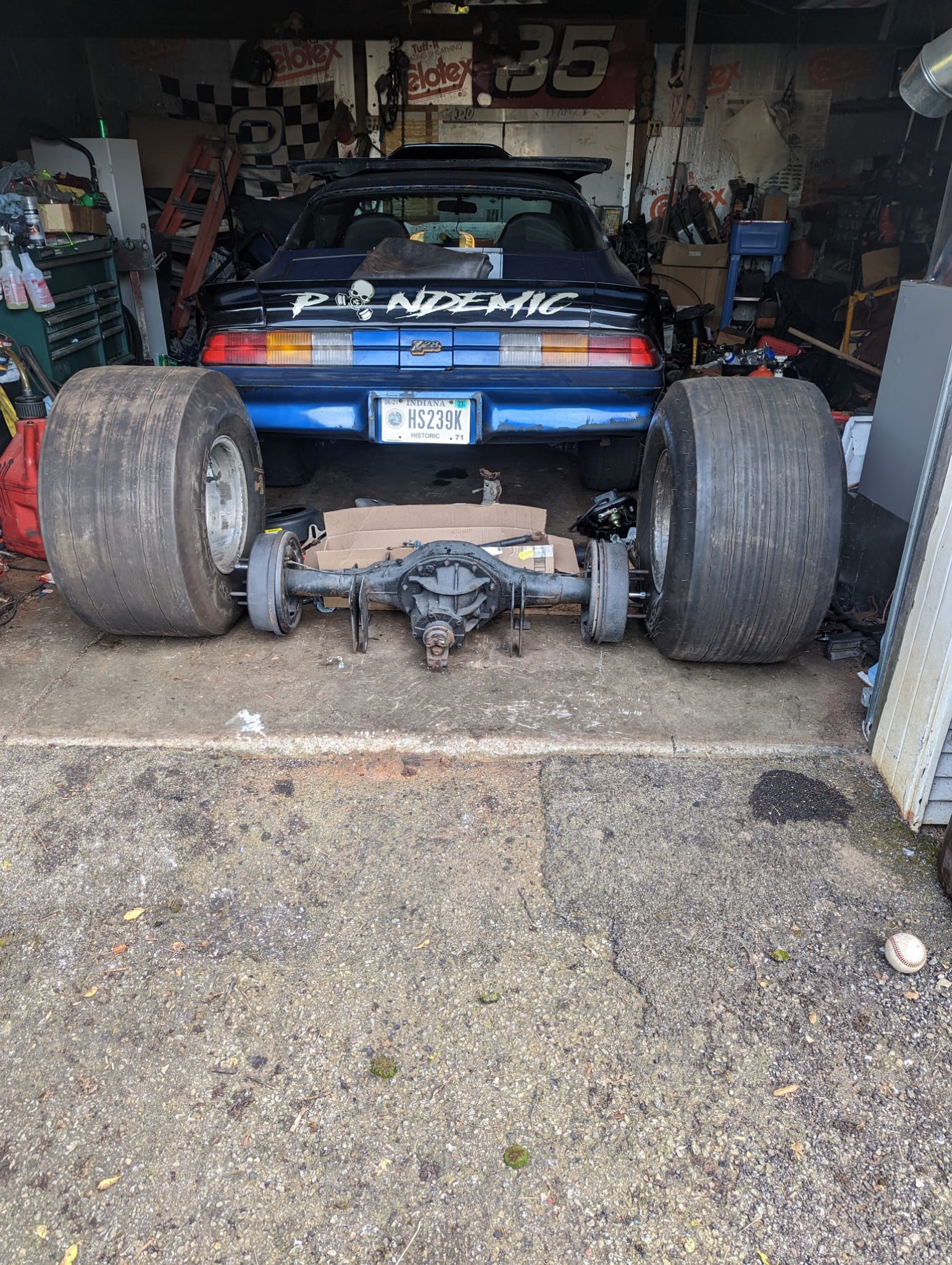 Rear view of a blue second-gen Camaro in a garage with massive drag slicks removed and a rear axle sitting on the floor.