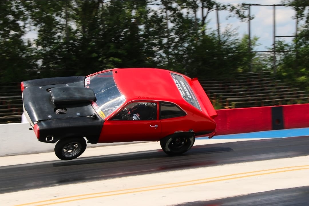 Red Team Chinto outlaw wheelstanding Pinto at extreme angle during a pass, front end high and car nearing rollover at the drag strip.
