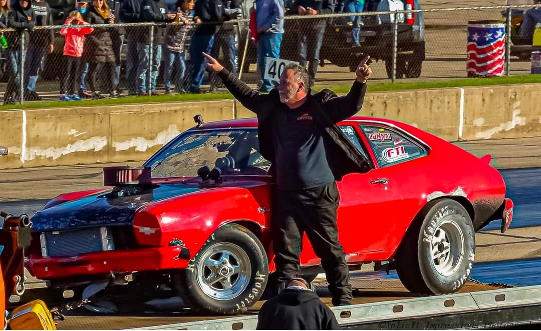 Red Team Chinto wheelstanding Pinto with the driver standing on the trailer celebrating a winning pass, crowd watching in the background.